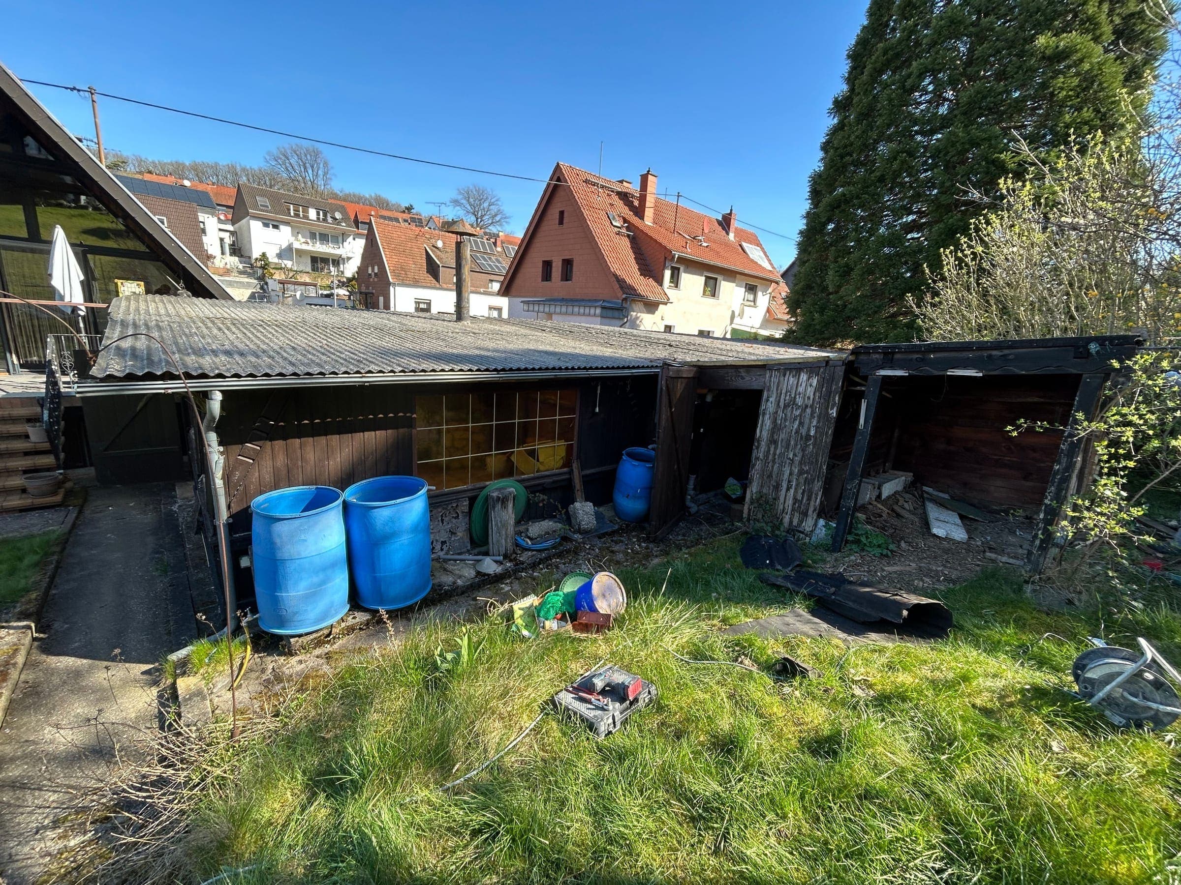 Baufällige Nebengebäudezeile mit Wellblechdach, Holzschuppen und abgestellten Fässern auf einem verwilderten Grundstück