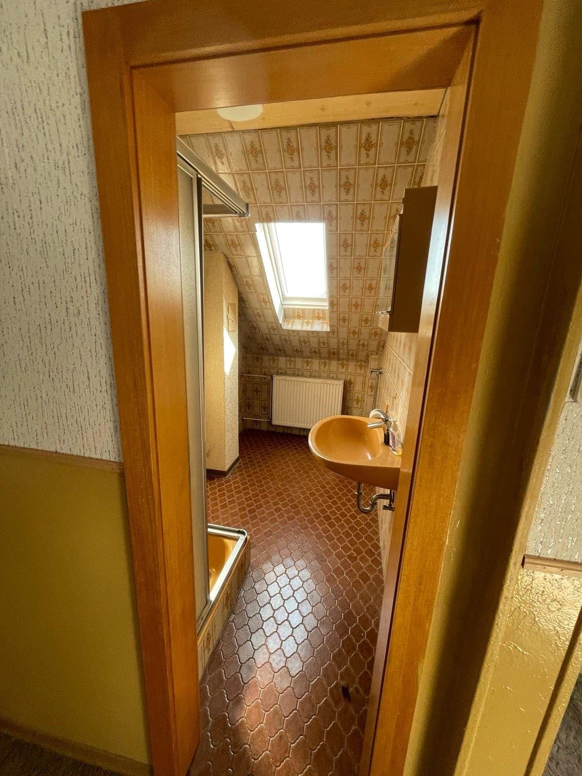 Blick aus dem Flur durch die offene Holztür in dasselbe 70er-Jahre-Bad mit orangefarbenem Waschbecken und braunen Sechseckfliesen