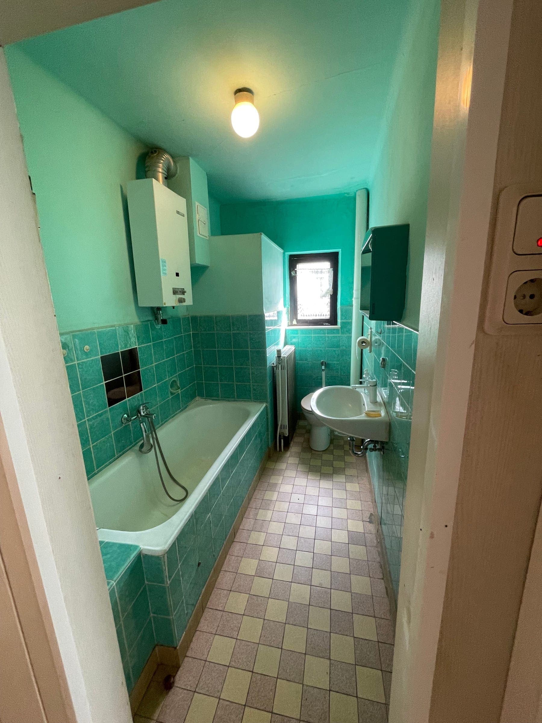 Altbaubad vor der Sanierung mit türkisen Wandfliesen, verkleideter Badewanne, Gastherme an der Wand und beige-braunen Bodenfliesen