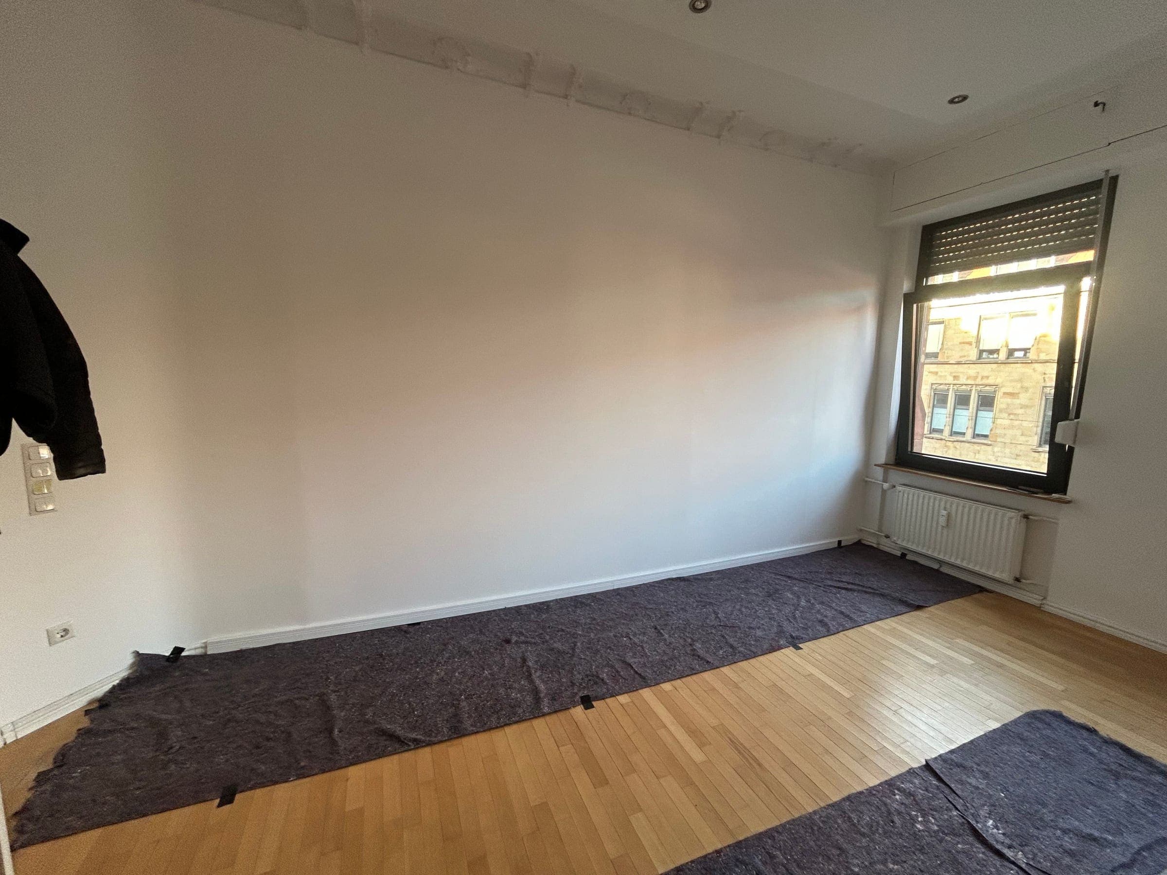 Fertig sanierter Altbauraum mit weißen Wänden, Eichenparkett unter Abdeckvlies und weißem Heizkörper unter dem hohen Fenster